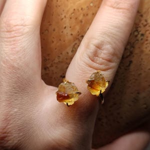 May include: A gold ring with two raw, orange-brown gemstones. The ring is on a finger.
