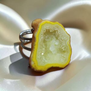Pode incluir: Um anel com uma grande fatia de geodo irregular, apresentando uma faixa externa amarelo-brilhante e um centro cristalino pálido. A pedra está montada numa banda de metal prateado composta por dois fios finos e paralelos ao redor do dedo.