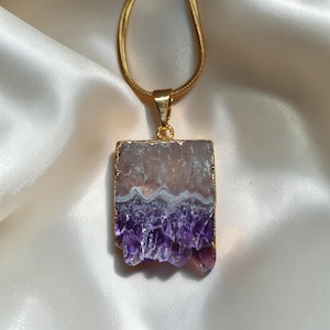 May include: A gold-toned snake chain necklace featuring a rectangular pendant with a raw amethyst crystal slice. The pendant displays a gradient from a translucent pale pinkish-white top section to a deep purple crystalline bottom edge.