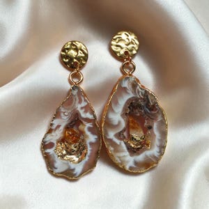 Puede incluir: Un par de pendientes colgantes con rodajas de ágata irregulares, translúcidas, de color marrón y blanco, con bordes dorados. Cada pendiente cuelga de un perno circular dorado martillado, con un pequeño cristal facetado de color ámbar en el centro.