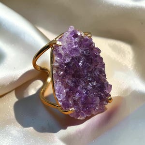May include: A gold-toned ring featuring a large, irregular, raw amethyst druzy stone as the centerpiece. The purple crystal cluster is held in place by a polished gold metal prong setting with a split, open-style band design.