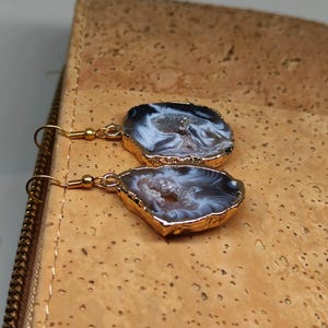 Puede incluir: Un par de pendientes de tono dorado con una piedra de &aacute;gata blanca y gris. Las piedras tienen una forma irregular y un aspecto natural y org&aacute;nico.