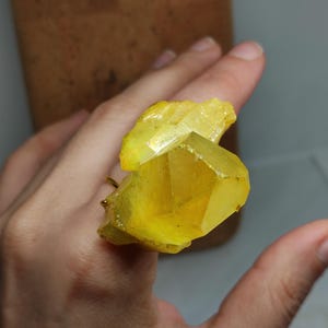 Könnte beinhalten: Ein Ring mit einem großen, rohen, hellgelben Kristallcluster als Herzstück. Der Kristall hat eine facettierte, geometrische Struktur mit durchscheinenden Kanten und einem lebendigen, gesättigten Gelbton. Er ist auf einem dünnen Metallring befestigt.
