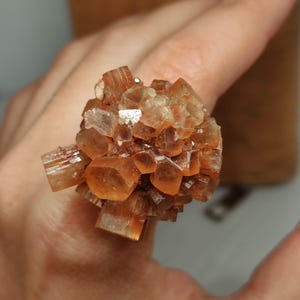 Può includere: Un anello con un gruppo di cristalli di aragonite marrone-arancio. I cristalli sono disposti in un motivo radiale, creando un design strutturato e accattivante. Un gioiello unico.