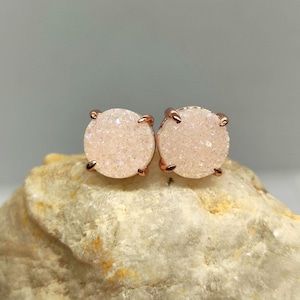 May include: A pair of round stud earrings with a light pink, sparkly druzy stone set in rose gold-colored metal. The earrings are displayed on a light-colored, textured rock surface.