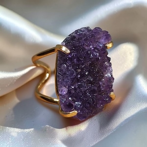 Op de afbeelding: Een goudkleurige ring met een grote, onregelmatige, dieppaarse amethist druzy steen als middelpunt. De steen heeft een getextureerd, kristallijn oppervlak dat op zijn plaats wordt gehouden door vier goudkleurige klauwen op een gespleten ringband.