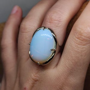 Könnte beinhalten: Ein Silberring mit einem großen, ovalen, opalisierenden Stein. Der Stein hat einen weichen, milchig-blauen Farbton mit subtilem Farbspiel. Die Fassung des Rings ist ein schlichtes, aber elegantes Design.