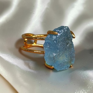 Op de afbeelding: Een goudkleurige ring met een grote, ruwe, lichtblauwe edelsteen in een klauwzetting. De ringband is opgebouwd uit twee parallelle dunne goudkleurige draden die samenkomen bij de centrale zetting, wat zorgt voor een open ontwerp.