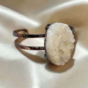 Könnte beinhalten: Ein Armreif mit einem dunkelmetallischen Band mit gehämmertem Muster und einem großen, ovalen, weißen Druzy-Edelstein als Mittelstück. Der Stein hat eine kristalline, strukturierte Oberfläche und ist fest auf dem offenen Metallrahmen montiert.