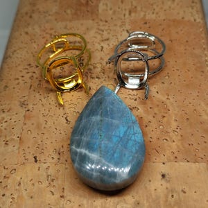 Peut inclure: Une pierre précieuse en labradorite bleue en forme de goutte avec des reflets irisés, placée devant deux montures de bague en métal, l'une dorée et l'autre argentée, chacune dotée d'un sertissage à griffes pour une pierre.