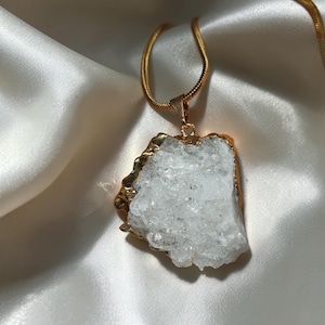 May include: A gold-toned snake chain necklace featuring a large, irregular, translucent white crystal pendant. The crystal has a rough, textured surface and is encased in a polished gold-coloured metal border that follows the natural shape of the stone.