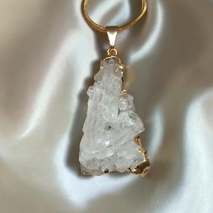 Op de afbeelding: Een heldere, grillige ruwe kwartskristal hanger hangend aan een goudkleurige slangenketting. Het kristal heeft een onregelmatig, veelzijdig oppervlak met een metalen goudkleurige rand die de steen aan een goudkleurig metalen oogje bevestigt.