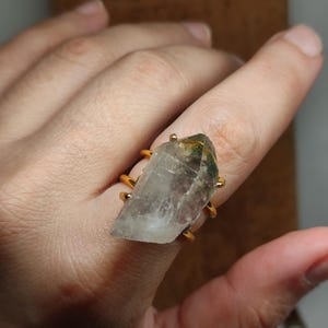 May include: A gold-toned metal ring featuring a large, raw, clear quartz crystal point as the centrepiece. The crystal has visible internal inclusions with green and brown hues, held securely in place by multiple thin gold-toned metal prongs.