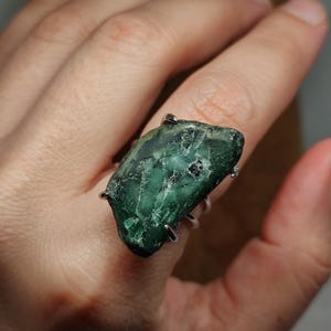 Pode incluir: Um anel com uma grande pedra preciosa bruta verde-escura, com inclusões brancas visíveis e uma textura rugosa e irregular. A pedra está fixada numa cravação de metal prateado com garras que envolve o dedo com uma banda fina e simples.