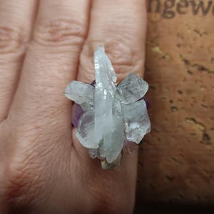 Puede incluir: Un anillo que presenta un grupo de cristales de cuarzo blanco crudo y translúcido sobre una base de piedras de amatista de color púrpura intenso. Las puntas de cristal naturales y dentadas están dispuestas verticalmente, creando un aspecto texturizado.