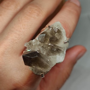 Puede incluir: Un anillo de cuarzo ahumado en bruto. El anillo presenta un gran cristal de cuarzo ahumado de forma irregular con una mezcla de tonos marrón claro y oscuro. El cristal está montado sobre una banda plateada con cuatro garras.