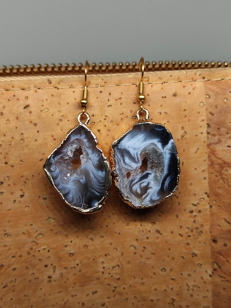 Puede incluir: Un par de pendientes de tono dorado con una rebanada de &aacute;gata blanca y gris con un borde dorado. Las rebanadas de &aacute;gata tienen una forma irregular y un aspecto natural y org&aacute;nico.