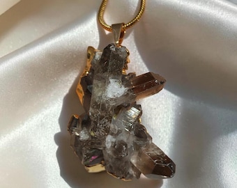 May include: A gold-toned snake chain necklace featuring a raw, dark brown smoky quartz crystal pendant. The crystal cluster has multiple pointed terminations and is encased in a gold-colored metal setting at the base, suspended from a gold bail.