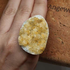 Könnte beinhalten: Ein Ring mit einer großen, unregelmäßigen, rohen Citrin-Geodenstufe mit durchscheinenden gelben und bernsteinfarbenen Kristallen. Der Stein hat einen weißen, strukturierten Außenrand und ist auf einer Ringschiene mit natürlicher Oberfläche.