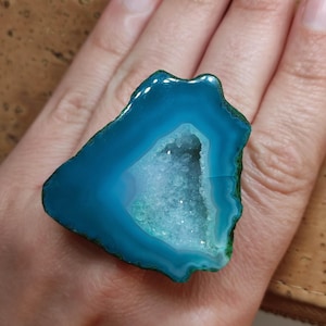 May include: A vibrant blue geode slice with a crystalline center. The polished stone has a natural, irregular shape and a band of teal around the edges. The geode is held in a hand, showcasing its color and texture.
