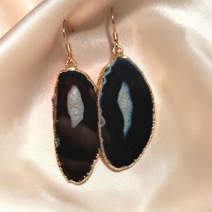 Puede incluir: Un par de pendientes colgantes con rodajas de ágata negra pulida y centros cristalinos blancos naturales. Cada piedra está engastada en un borde de metal dorado fino y suspendida de un gancho a juego para orejas perforadas.