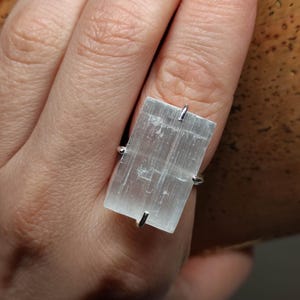 Puede incluir: Un anillo de plata con un cristal de selenita rectangular transparente. El cristal tiene una textura estriada.