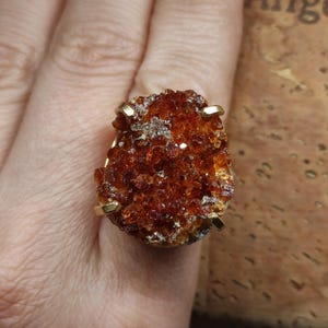 Könnte beinhalten: Ein Ring mit einem großen, rohen, bernsteinfarbenen Druzy-Edelstein mit einer texturierten, kristallinen Oberfläche. Der Stein wird von vier goldfarbenen Metallkrallen gehalten, die das Cluster fixieren und ein markantes, organisches Mittelstück bilden.