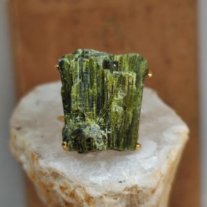 Op de afbeelding: Een ring met een ruw, verticaal gestreept olijfgroen toermalijnkristal in een goudkleurige metalen zetting. De edelsteen heeft een natuurlijke, ruwe textuur met diepgroene en geelgroene kleurvariaties over de gehele steen.