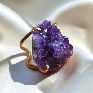 Könnte beinhalten: Ein goldfarbener Ring mit einer großen, rohen, tiefvioletten Amethyst-Geoden-Gruppe als Herzstück. Der Stein wird von vier dünnen Metallkrallen gehalten, und die Ringschiene hat ein wellenförmiges, offenes Design, das sich um den Kristall legt.