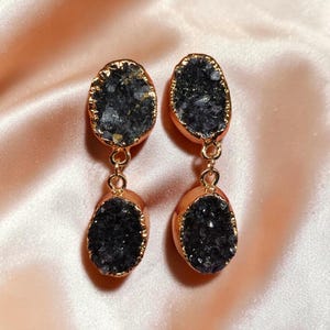 May include: A pair of dangle earrings featuring a gold-colored frame with a dark, textured stone center. The earrings have a double-stone design, with a smaller stone hanging below a larger one. The background is a soft, peach-colored fabric.