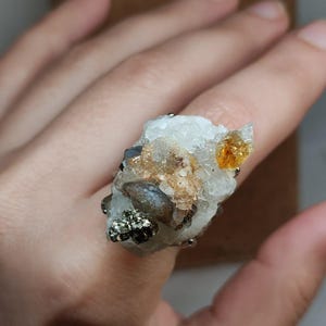 Puede incluir: Un anillo único con un grupo de cristales y minerales naturales. El anillo presenta una mezcla de elementos blancos, dorados y metálicos oscuros, con una prominente formación de cristal texturizado. El diseño es orgánico y llamativo.