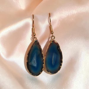 Puede incluir: Un par de pendientes colgantes con rodajas de ágata azul pulidas en forma de lágrima con bandas naturales. Cada piedra está engastada en un fino borde de metal dorado y suspendida de un gancho dorado a juego para orejas perforadas.