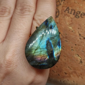 Könnte beinhalten: Ein tropfenförmiger Labradorit-Edelsteinring mit irisierenden Blitzen in Blau, Grün und Gold in einem dunkelgrauen Stein. Der polierte Cabochon ist in eine goldfarbene Metallfassung eingefasst und zeigt natürliche Einschlüsse.