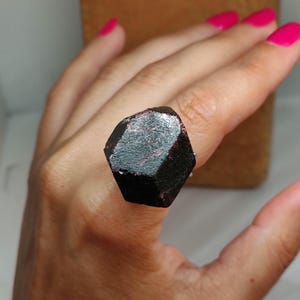 May include: A dark, faceted gemstone ring. The large, geometric stone is a deep, almost black color with hints of red. The ring is set on a silver band. The background is a neutral tan color.