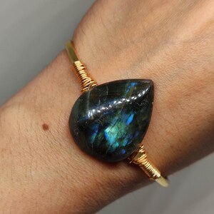 May include: A gold-coloured wire bracelet with a teardrop-shaped labradorite stone. The stone has iridescent blue and green flashes. The wire is wrapped around the stone and the bracelet's band.