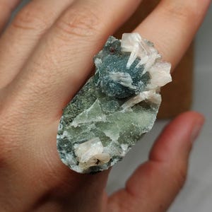 May include: A unique ring featuring a natural mineral specimen. The ring showcases a rough-cut, green and blue stone with crystalline formations. The stone is held in place by a hand.
