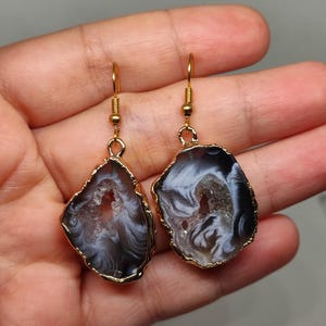 Puede incluir: Un par de pendientes de tono dorado con una rebanada de &aacute;gata negra y blanca con un centro cristalino. Los pendientes tienen un dise&ntilde;o colgante.