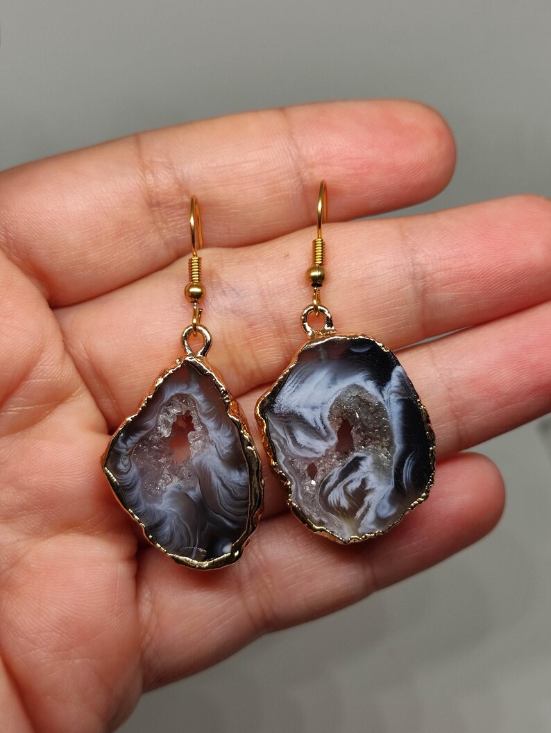 Puede incluir: Un par de pendientes de tono dorado con una rebanada de &aacute;gata negra y blanca con un centro cristalino. Los pendientes tienen un dise&ntilde;o colgante.