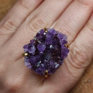 May include: A gold ring with a large, raw amethyst crystal. The amethyst is a deep purple colour with a textured surface. The ring's setting has four gold prongs holding the crystal in place.