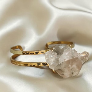 Puede incluir: Un brazalete tipo puño de tono dorado con un diseño de doble banda y textura martillada. Un cristal de cuarzo bruto, grande y transparente, está montado de forma segura en el centro, actuando como el punto focal de este accesorio de metal.