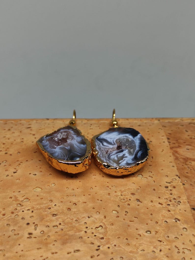 Puede incluir: Un par de pendientes de &aacute;gata chapados en oro con una piedra de &aacute;gata blanca y gris. Los pendientes tienen forma de semic&iacute;rculo y se cierran con un gancho.