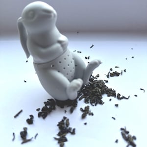 May include: A white ceramic bunny tea infuser with a perforated belly, surrounded by loose tea leaves. The bunny is sitting, with its ears and legs extended. A charming and functional kitchen accessory.