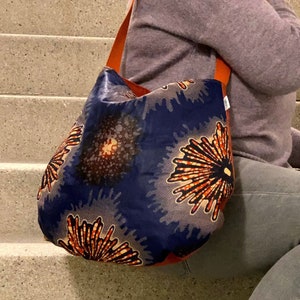 Op de afbeelding: Een blauwe en oranje gepatroneerde tote bag met bruine leren hengsels. De tas wordt over de schouder gedragen door iemand die een grijze jeans en zwarte enkellaarsjes draagt.