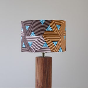 Op de afbeelding: Een houten tafellamp met een cilindrische basis en een lampenkap met een patroon. De lampenkap heeft een geometrisch ontwerp met bruine, oranje en blauwe driehoeken.