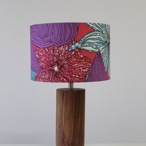 Könnte beinhalten: Eine Tischlampe aus Holz mit einem zylindrischen Sockel und einem bunten Lampenschirm mit floralem Muster. Der Lampenschirm zeigt rosa, lila und blaue Blumen auf rotem Hintergrund.