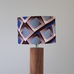 Op de afbeelding: Houten tafellamp met een cilindrische voet en een patroon lampenkap. De lampenkap heeft een geometrisch ontwerp met blauwe, bruine en roze ruitvormen. De lamp is een decoratieve verlichtingsarmatuur.