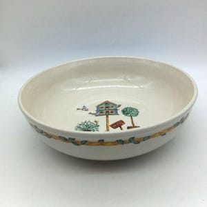 May include: Off-white ceramic serving bowl with a birdhouse and tree design. The bowl has a yellow and green leaf border. The interior features a birdhouse, a tree, and a sign. The bowl is suitable for serving salads, pasta, or other dishes.