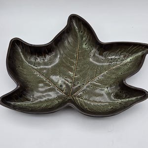 May include: A ceramic dish shaped like a large maple leaf with a glossy finish. The surface features a mottled blend of dark brown and sage green glaze, with embossed vein details radiating from the center to the edges of the leaf-shaped bowl.