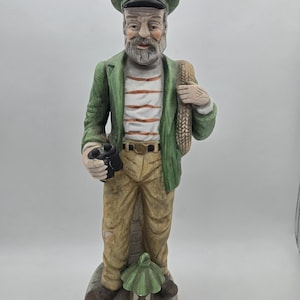 May include: A ceramic figurine of an elderly sailor, wearing a green jacket, striped shirt, and khaki pants. He holds binoculars and a rope over his shoulder. The figurine stands on a stone base with a lantern.