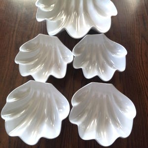 May include: A set of five white, shell-shaped serving dishes. The set includes one large dish and four smaller dishes, all with a glossy finish. The dishes are arranged on a dark wooden surface.